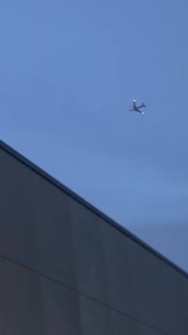 The video shows a gray building with a flat roof and a plane flying overhead. The sky is light blue. The plane has lights on. The video pans up to follow the plane.