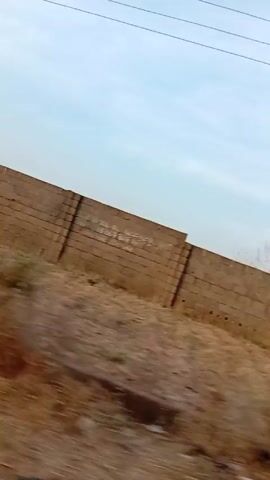 The video appears to be taken from a moving vehicle, likely a car, driving on a paved road next to a wall. The wall is made of concrete blocks and extends along the side of the road. The area between the road and the wall is mostly dry, reddish-brown soil with some sparse vegetation. Beyond the wall, trees can be seen in the distance. The sky is overcast. The perspective and speed of the video suggest that the camera is inside a moving vehicle.