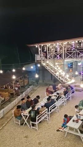 The video shows an outdoor restaurant with multiple levels at night. The lower level has white picnic tables and benches set up on a gravel ground, with people sitting at them and eating. There are string lights illuminating the area. A staircase leads up to a second level, which also has tables and people. The second level appears to be an enclosed structure with a bar area. Further back, there is a road with parked motorcycles and several small food stalls or shops. The overall atmosphere is casual and illuminated by artificial lights.