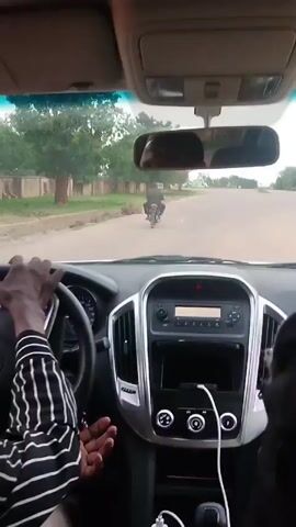 Here's a detailed description of the video:

**Scene:** The video is shot from the driver's perspective inside a car. The driver is driving down a paved road lined with trees and fences. 

**People:** The main visible person is the driver, an adult with dark skin, wearing a black and white striped long-sleeved shirt. Their hands are on the steering wheel.  

**Objects:**
*   **Car:** The car has a gray and black interior. The dashboard features a digital display (partially visible) on the steering wheel, a radio and what appears to be an infotainment system in the center, and climate control knobs below. A white cable is plugged into a USB port in the car's center console.
*   **Road:** The road is gray asphalt with a white line on the shoulder, and there's another person riding a motorbike further down the road.
*   **Environment:** Green trees and low fences can be seen along the roadside.

**Actions/Interactions:** The driver is operating the vehicle, gripping the steering wheel. The car appears to be moving forward.

**Environment:** The sky is overcast or slightly hazy. The surroundings seem to be a suburban or semi-rural area.