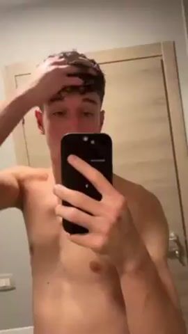 A shirtless young man stands in a bathroom taking a selfie video. He is fair-skinned, with short brown curly hair, and brown eyes. He runs his fingers through his hair and looks at the camera.  Behind him is a light-colored wooden door and a gray wall with a light switch.  A small box sits atop the door frame.