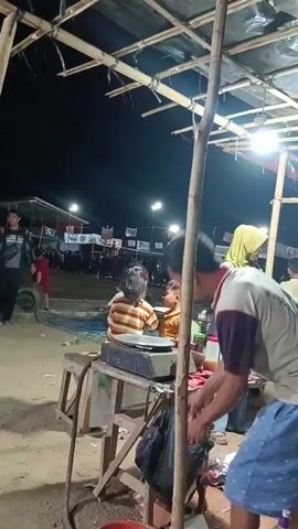 The video shows a nighttime outdoor scene, possibly a market or fair, with several people present. In the foreground, a person stands facing away from the camera, wearing a light-colored t-shirt with a design and patterned shorts. Next to them, a wooden table holds a dark-colored flat-top cooking appliance. Two children and an adult are seated or leaning over the table, appearing to be interacting with the cooking surface. To the left, a young girl in a striped shirt and green shorts walks towards the center, holding something in her hand. Further in the background, more people are visible, some walking and others gathered, with a structure that looks like a stage or seating area adorned with banners. Lights illuminate the area, indicating it is dark outside. The ground appears to be dirt or gravel. The overall atmosphere suggests a lively event taking place at night.