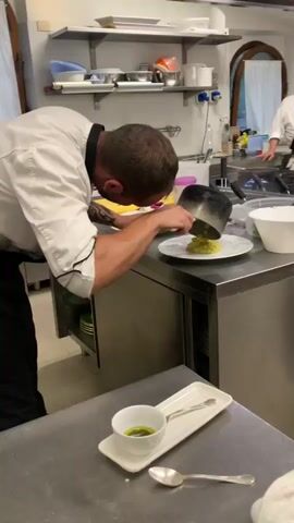 A chef in a white chef's coat and black apron is plating food. He is leaning over a stainless-steel counter, using a blue spatula to scrape food from a silver pot onto a white plate. The food appears to be a yellow, creamy dish, possibly risotto. In the foreground, a small white bowl with green liquid and a spoon on a rectangular white dish are visible. Another white bowl and some kitchen equipment are on the counter behind the chef. Another person, also wearing a chef's coat, is visible in the background to the right. The environment appears to be a professional kitchen.