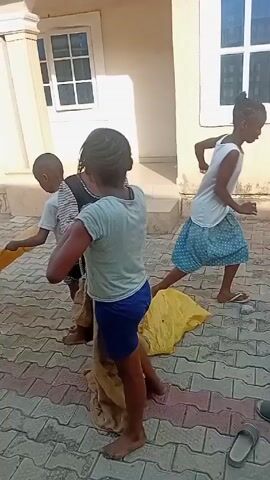 Three young children, two girls and one boy, are on a red and gray brick paved surface in front of a light-colored building. The girls are standing, while the boy is sitting on a tan sack or cloth. One girl wears a light blue t-shirt and blue shorts, while the other wears a long-sleeved striped dress. The boy is wearing a t-shirt and shorts. The girl in the dress is holding up a brown sack, while the other girl is holding up a yellow sack. There is a discarded sandal on the brick paving. The building has white window frames and what appear to be classical-style white columns.