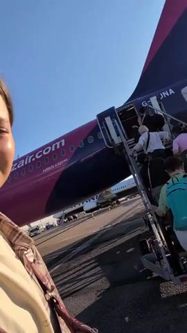 The video shows a group of passengers boarding an airplane using a mobile staircase. The airplane is predominantly pink and purple with "wizzair.com" written on its side. The tail of the plane is visible, also pink. The sky is clear and blue. The first segment shows people ascending the stairs, carrying luggage. The subsequent segments focus on a young woman with her hair pulled back, wearing a plaid shirt over a yellow t-shirt and a beaded necklace. She is looking at the camera with a tired or resigned expression, occasionally blinking and making facial gestures. The background in these segments shows the airplane and the sky. The scene appears to be at an airport tarmac during daylight.