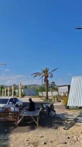 The video captures an outdoor scene with a bright blue sky. In the foreground, there is a small food cart and a few motorcycles parked on a paved area. A person is seated on a bamboo mat. Palm trees are visible, one of which is prominent in the center. In the background, there are buildings with architectural features reminiscent of the Kaaba. Large white umbrella-like structures, possibly for shade, are also present. The overall environment appears to be a public space, possibly a religious site or a tourist attraction.