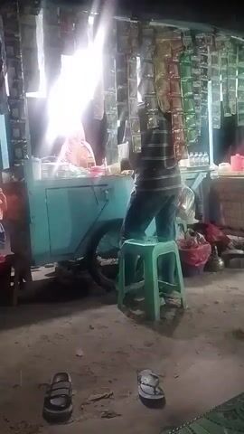 The video shows a nighttime scene in what appears to be a small outdoor market or street vendor area. A person is standing on a green stool behind a turquoise cart, which is illuminated by a bright light. The cart is laden with various items, including what look like packaged goods hanging in rows. In front of the cart, a person in a striped shirt and jeans is interacting with the vendor. Several children are also present. One child, wearing an orange shirt and blue shorts, stands near the cart looking towards it. Another child, partially visible in the foreground, is facing away from the camera, with their hand near their face. Other adults and children are in the background, some walking away from the cart. The ground is dirt, and sandals are visible on the ground. The overall atmosphere suggests a casual, possibly bustling, evening gathering.