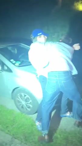 At night, two young men are grappling near the front of a silver car, lit dimly by the car’s headlights and an unseen light source. One man wears a blue baseball cap, light blue button-down shirt, dark pants, and brown shoes with patterned socks visible. The other wears a striped button-down shirt, dark jeans, and dark shoes with white stripes. They are pushing and shoving each other, with the man in the baseball cap appearing to strike the other. A hand reaches into the frame briefly, seeming to try and break them up. The background is dark, with some vegetation visible and a tree in the distance.