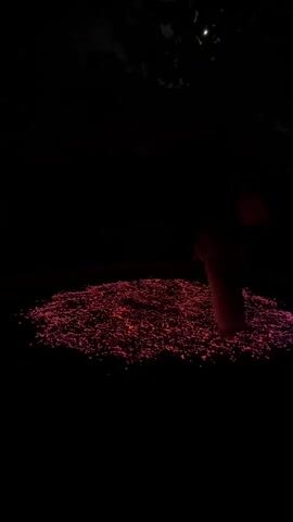 A person in a long, flowing orange garment stands on a circular patch of glowing pink and red particles in a dark outdoor setting. Above the scene, silhouetted tree branches are visible against a dark sky with a faint crescent moon. The person is partially obscured by darkness and the glow of the particles, appearing to be performing some kind of ritualistic movement or dance.