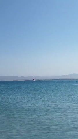 The video shows a wide shot of a calm, blue-green body of water under a clear, light blue sky. In the distance, a small sailboat with white and red stripes on its sail is visible, moving across the water. Farther back, a line of hazy mountains or distant land masses stretches across the horizon. The water is mostly smooth with small ripples, and a faint dark line, possibly buoys or a distant structure, can be seen in the mid-ground. To the right, a dark, flat object floats on the water. The scene suggests a bright, sunny day in a coastal area, likely in Safaga, Egypt, as per the location context. A child's voice is heard calling out "Papa" at the beginning of the video.