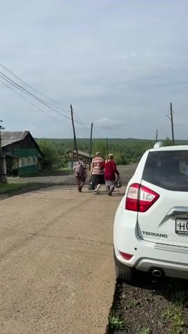 Two women and a third person are walking away from the camera on a dirt road. The woman in the middle is wearing a striped shirt and shorts, and she is carrying a bag. The woman on the right is wearing a red shirt and dark pants, and she appears to be using a cane. The person on the left is wearing a patterned shirt and dark pants, and they are carrying a backpack. A white SUV is parked on the right side of the road, partially obscuring the view of the people. In the background, there are wooden houses, trees, and utility poles under a cloudy sky.