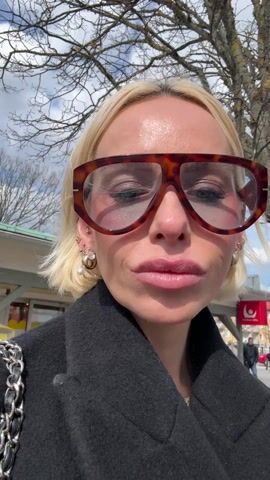 The video shows a close-up shot of a fair-skinned blonde woman wearing oversized tortoise-shell glasses, a black coat, and various earrings on her left ear. She is facing the camera and smiling. She raises her right index finger up to her mouth while announcing she's about to use her Collective Memory debit card to purchase something. The background shows a slightly overcast sky, bare tree branches, a building, and a sign, which is partially obscured. A person in a dark coat is walking in the background.