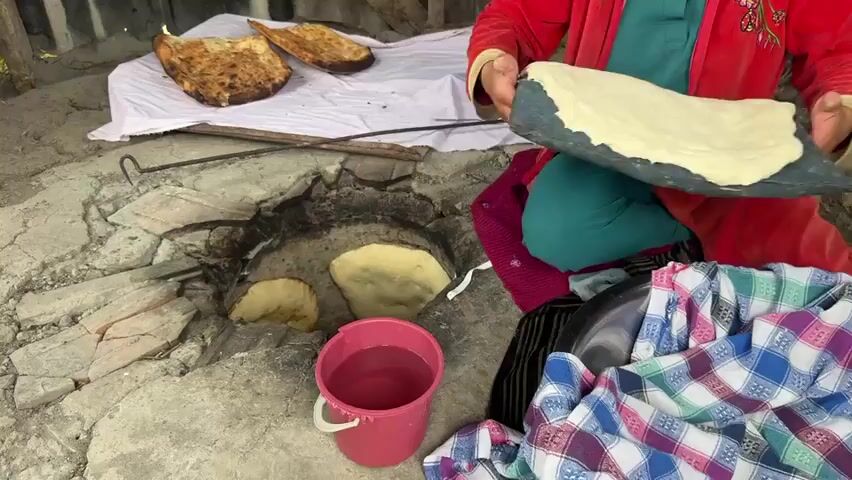 A person in a red jacket and teal garment is cooking flatbread in a tandoor oven. They are holding a flattened piece of dough on a dark paddle, about to place it inside the hot oven. Inside the tandoor, several pieces of already baked flatbread are visible, some browning. Two pieces of golden-brown, baked flatbread are laid out on a white cloth to the left of the oven. A pink bucket with water is positioned in front of the oven. A metal bowl with patterned cloth is to the right. The tandoor itself is a circular pit dug into the ground, with broken bricks and concrete surrounding it. The background suggests an outdoor setting, possibly a rural or traditional area.