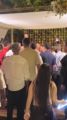 A crowd of people are gathered under a canopy adorned with white fabric and string lights. The people are predominantly men wearing white shirts and kippahs, indicating a possible religious event. Some individuals are holding up phones, appearing to record or take pictures. The overall atmosphere seems festive and celebratory, with people interacting and engaged in the event. The background shows foliage and trees, suggesting an outdoor setting.