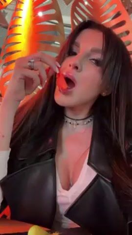 The video shows a woman with long, dark hair and a black leather jacket, eating what appears to be a red-colored food item. She is wearing a white shirt with a V-neck, exposing a tattoo on her chest. She has a choker necklace with a cross pendant and rings on her left hand. The background includes an orange, decorative structure and what looks like a table with a lemon and other items. The lighting is warm, with orange hues dominating the scene. The focus is on the woman as she puts the food in her mouth and chews.