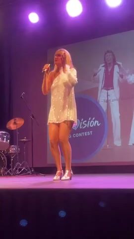 A woman with blonde hair is standing on a stage, holding a microphone and singing. She is wearing a sparkly white one-shoulder dress and silver heels. The stage has purple lighting, and a drum set is visible to the left. Behind the woman, a large screen displays the Eurovision Song Contest logo and an image of a man in a white suit. The woman is looking upwards and to her right, appearing to be in the middle of a performance.