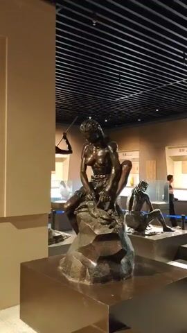 The video opens with a wide shot of a museum exhibit. The ceiling is a dark, grid-like structure with lights. In the foreground, a dark brown sculpture of a human figure is visible, crouched on a pedestal. The camera pans slightly to the right, revealing more of the exhibit space. In the mid-ground, another dark brown sculpture of a seated human figure is present, also on a pedestal. Behind this, several display cases with glass tops are arranged, some with objects inside. Further in the background, another full-body sculpture of a human figure is seen, standing with a bow and arrow drawn, seemingly aiming. This figure is also dark brown. A person, wearing a dark shirt and pants, stands near the bow-and-arrow sculpture, looking towards it. To the left, another person, also in dark clothing, is partially visible near a display case. The lighting in the room is subdued, with spotlights illuminating the sculptures and display cases. The walls are a neutral beige color. The camera then briefly pans back towards the crouching sculpture and then moves to show more of the exhibit area, including additional display cases and a person walking in the distance. The video ends with a shot showing the standing bow-and-arrow sculpture prominently in the foreground with a person in the background.