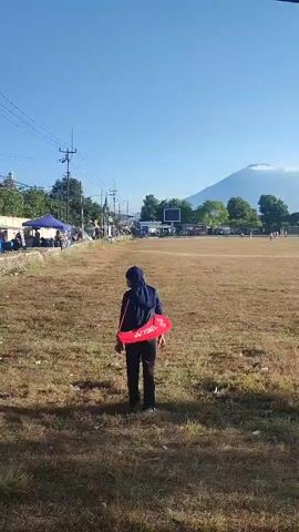 The video shows a person wearing a blue hoodie, black pants, and a blue headscarf walking across a dry, grassy field. They are carrying a red bag over their right shoulder. In the background, there is a mountain with a cloud on top, a soccer field with players, a few buildings, and some trees. The sky is clear and blue. The lighting suggests it is daytime, likely morning or afternoon.