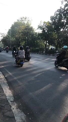 The media file shows a street scene in Gelam, Indonesia, with several people riding motorbikes. The sun is low in the sky, suggesting it is either early morning or late afternoon, and the weather appears to be clear. The road is paved and has a marked lane. Lush green trees line the sides of the road, with some buildings visible in the background.

The main subjects are the people on the motorbikes, most of whom are wearing helmets for safety. The person in the foreground is wearing a red jacket and appears to be riding a dark-colored motorbike. Other individuals on motorbikes are seen in the background, traveling in the same direction. The overall atmosphere seems calm and ordinary, depicting daily life in a Southeast Asian urban or suburban environment. There are no visible signs or text in the media file.