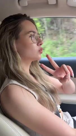 The video shows a woman in the passenger seat of a car, giving a peace sign and a kiss to the camera. The camera then shifts to the driver's side showing the steering wheel and then the dashboard. The infotainment screen displays a map and music playing. The car is traveling on a paved road surrounded by lush green trees. The sky is overcast.