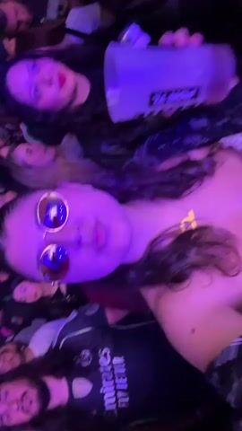 A woman with long dark curly hair and round sunglasses is holding a large white cup with a black logo on it and taking a sip from it. She is surrounded by other people in what appears to be a crowded indoor venue with purple lighting. She is wearing a plaid shirt. The cup has the word "PORTA" visible on it.