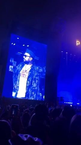 The video shows a concert crowd from a low-angle perspective, looking up towards a large screen displaying a performer. The performer, wearing sunglasses and a cap, is illuminated on the screen, superimposed over what appears to be a collage of smaller images. Behind the screen, the stage is bathed in intense blue light. Above the stage, large, illuminated letters spell out "PARK LIVE." The crowd is a dark mass of silhouetted figures, many holding up their phones to record or photograph the performance. The overall ambiance is that of a large-scale live music event, characterized by bright stage lighting and enthusiastic audience participation.