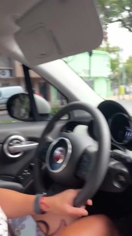 The video shows a woman driving a car. She is looking to the side and has one hand on the steering wheel, the other appears to be holding a cell phone on her lap. Her hair is pulled back and she is wearing earrings. The car's interior is dark with a Fiat steering wheel logo, and the exterior environment is visible through the windows. Buildings and a motorcycle can be seen outside.