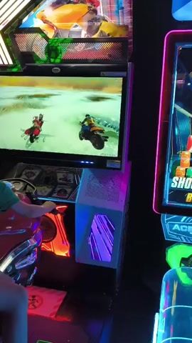 The video shows two children playing an arcade game that appears to be a motorcycle racing game. The game features bright graphics and colorful flashing lights. The children are sitting on a simulated motorcycle controller, which is attached to a large screen displaying the game. One child is in the front, steering the motorcycle, and the other child is sitting behind them. The game screen shows a race in progress, with multiple motorcycles on a track. The environment is dimly lit, typical of an arcade, with the game's lighting being the primary source of illumination. The game board above the screen displays a stylized logo with the letters "EX". To the right of the racing game, another arcade game with neon lights and a display showing "SHOOT BOX" is partially visible.