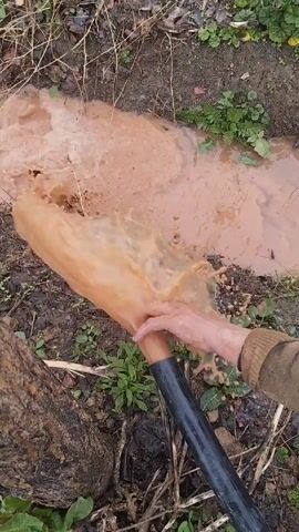 The scene features a person in a brown sweater holding a black and brown hose, from which a stream of muddy, brown water is flowing. The water forms a small pool on the ground, which is covered in dirt, dead leaves, and some green plants. A portion of a tree trunk is visible on the left side of the frame. The person is directing the flow of water into the existing pool on the ground.
