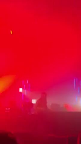 A DJ is performing on stage in a club or concert venue. The lighting alternates between red and blue, with some spotlights and blue vertical light bars. The stage has a "TRANS MUSICALE" logo. The DJ is facing the equipment, mixing music. There are silhouettes of audience members in the foreground. There is also fog or haze on the stage.