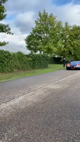 The video shows a black and orange Bugatti Veyron driving on a gray asphalt road with green trees and bushes on either side. The car has orange rims and is emblazoned with the number "84" in a white circle. A man wearing a dark jacket is visible standing near the car as it approaches. The video follows the car as it turns and drives away. The background shows a cloudy sky with glimpses of blue. Other people are visible taking pictures.