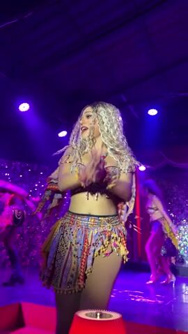The video shows a female dancer with long, curly blonde hair on a stage. She is wearing a colorful, patterned outfit consisting of a bra top and a short skirt with fringe. She is dancing and smiling, with her arms outstretched. Other dancers, also in colorful costumes, are visible in the background. The stage is lit with purple and red lights, and there is a crowd of people in the audience, many holding up lights. The environment appears to be a nightclub or performance venue.