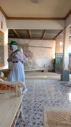The video shows two individuals in prayer within an interior space. The environment appears to be a courtyard or an open-air area adjacent to a building. The walls are a mix of concrete and corrugated metal, with some areas showing signs of wear and tear. One individual is in the foreground, kneeling with their forehead touching the ground. They are wearing a white garment and a green headscarf. Another individual is further back, also in prayer, standing and then bowing. This individual is wearing a white garment and a dark head covering. To the right, a decorative white wooden structure with floral embellishments is visible. There is also a large, yellow vase-like object and a fan in the background. The lighting suggests it is daytime.
