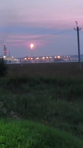 The video begins with a view of a field at dusk. In the background, a cluster of industrial buildings with lights can be seen, and a tall flare stack is burning, emitting a bright flame. A power line pole with wires is visible in the foreground to the right. The sky is a gradient of purple and pink, indicating twilight. The camera is stationary, capturing the scene without any significant movement. The overall atmosphere is one of industrial activity set against a natural, fading light.