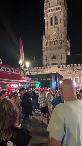 The video begins with a view of a crowded outdoor event at night. A tall, stone building with a clock tower is visible in the background. In the foreground, a crowd of people are gathered, facing a stage set up in front of the building. On the stage, a group of people in red shirts are performing, illuminated by green and blue stage lights. A banner with the word "Jupiler" is visible on a red structure to the left of the stage. The overall atmosphere appears festive. The camera angle shifts slightly, offering a closer view of the stage performance and the audience. The video concludes with the performance continuing and the crowd remaining engaged.