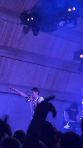 The video captures a live outdoor concert at night. In the foreground, a crowd of people is visible, many with their arms raised. A performer is on stage, illuminated by blue lights, singing into a microphone and gesturing with their free hand. The stage has a wooden structure above it, with lights mounted on it. The background features trees and a dark sky, with some tents and string lights suggesting a festival or event setting. The lighting is predominantly blue, with some hints of other colors, creating a vibrant atmosphere. The crowd appears engaged and enthusiastic.