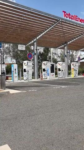 A wide shot shows a row of electric vehicle charging stations under a covered canopy at a TotalEnergies station. The charging stations are white and silver with digital screens. Several cars are parked at the stations, and a white van is visible in the background. The sky is blue with some clouds.