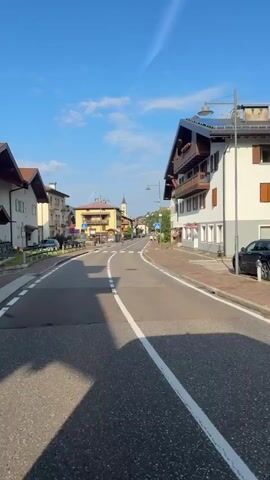 The video shows a view from a moving vehicle on a paved road. The road has white lane markings. On the right side of the road are buildings with balconies and solar panels. There are parked cars, including a black SUV and a white van. A red cylindrical object is visible on the sidewalk. On the left side of the road are more buildings and some greenery. In the distance, a town with a church steeple is visible. The sky is blue with a few wispy clouds. There are no people clearly visible in the foreground, but there may be individuals on bicycles or walking in the distance.
