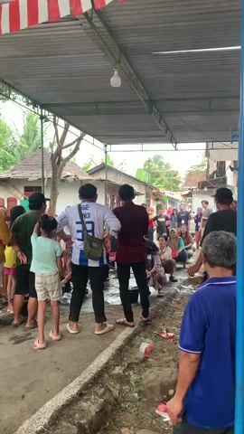 A group of people are gathered under a metal awning decorated with red and white striped fabric. The awning covers a dirt and gravel path with some puddles. In the foreground, a man in a blue shirt stands with his back to the camera. Behind him, a person in a maroon shirt and another in a white shirt with blue stripes are standing. To the left, several people, including children, are also standing or sitting. Further back, more people are visible, some sitting on the ground and others standing. Houses are visible in the background. Some trash is scattered on the ground. The overall impression is a community gathering or event taking place outdoors.