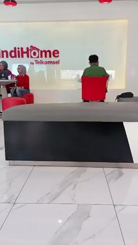 Here is a concise description of the scene in the video:

The video depicts the interior of an IndiHome by Telkomsel store. The store features modern decor with white marble floors and a large glass wall displaying the IndiHome logo. There are several red chairs and stools around small white tables, where individuals are seated. Some are working on laptops, while others appear to be consulting with employees of the store, identifiable by their red uniforms. The lighting is bright, and the overall atmosphere is clean and professional.