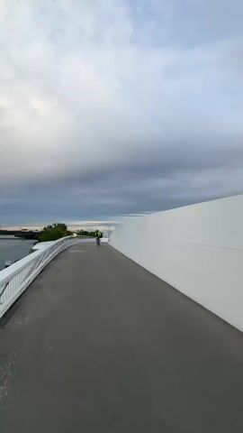 A person cycles away from the camera on a modern, white bridge over a river in Raunheim, Germany. The bridge features a sleek, curved white wall on the right and a white metal railing overlooking the water on the left. The cyclist, wearing a bright yellow vest, is visible in the distance on the grey paved path. To the left, a calm river flows, with green trees lining the distant banks. Another bridge is faintly visible in the far distance over the water. The sky is overcast with a mix of light and dark clouds, suggesting either early morning or late afternoon. The overall atmosphere is quiet and serene.