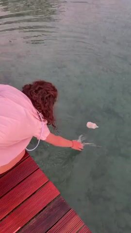 A person with curly brown hair, wearing a pink shirt and denim shorts, is kneeling on a wooden deck next to a body of clear water. The person is reaching into the water with their right hand. In the water, about a meter away from the deck, a white object, possibly a baby bottle, is floating. The person appears to be trying to retrieve the object from the water. The water is calm with ripples visible. The lighting suggests it is either early morning or late evening.