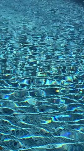 The video shows a close-up of rippling water in a swimming pool. The water is a bright blue color, and sunlight is reflecting off the surface, creating shimmering patterns. The bottom of the pool appears to be tiled with small, dark blue squares. There are no people or other objects visible in the video. The scene is calm and tranquil.