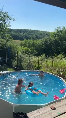 Three young people are playing in a backyard swimming pool on a sunny day. The pool is round with a white liner and a metal frame. The water is clear and blue, reflecting the sunlight. The surrounding area is lush with green trees and foliage, and a rolling hill can be seen in the background. A wooden deck is in the foreground of the image.

In the pool, one person, wearing a dark hat, is in the water on the far side, appearing to toss a blue ball. Another person, on the left, is partially submerged and facing the person in the blue inner tube. The third person is in the center, lying on their back in a bright blue inner tube, wearing goggles. This person is looking towards the person on the left. Two pink flamingo floaties are visible in the water near the edge of the pool.