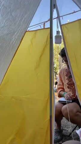 Inside a tent with yellow fabric walls, a person wearing an orange shirt and black shorts is sitting. They are holding a notebook and a pen, and appear to be writing. Outside the tent, several other people are visible, walking around. One person in a white shirt and black shorts is holding what appears to be a tennis racket. The environment outside appears to be outdoors with trees and sunlight.