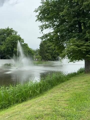 The video shows a body of water with fountains spraying water. The water is dark, and the fountains are creating white plumes of spray. The water is surrounded by lush green grass and trees. The sky is overcast, suggesting a cloudy day. The scene appears to be a park or a garden with a pond or lake. The presence of multiple fountains indicates a designed landscape. There are no people visible in the video. The overall impression is one of a serene, natural setting enhanced by man-made water features.