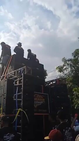 The video shows a crowd of people gathered around a large stack of speakers at an outdoor event, likely a street party. The speaker system, labeled "SAK MUNINE AUDIO," is set up on a trailer or platform, with several individuals standing on top, possibly monitoring or adjusting the equipment. The crowd consists of people of various ages, including children and young adults, many wearing matching black t-shirts with a graphic design on the back. Some people are holding cups, suggesting they are drinking. The environment appears to be a residential street, with houses and trees visible in the background. The sky is bright with some clouds. The camera is positioned within the crowd, capturing the scene from a ground-level perspective. The overall atmosphere seems lively and festive, with music likely being played through the sound system.