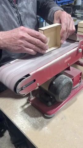 A person is using a belt sander to smooth the edge of a small wooden box. They hold the box against the moving belt and apply pressure. They then examine the box before placing it on a stack of similar wooden boxes. The belt sander is red, with a black motor. The person is wearing a dark jacket. The work area is cluttered with tools and materials.