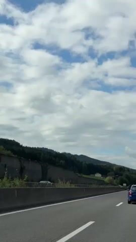 The video shows a view from inside a car traveling on a highway. The camera is pointed forward and slightly to the right. The highway has a concrete barrier on the left side, and a green, tree-covered hillside beyond it. The sky is blue with white, fluffy clouds. Several cars are visible on the highway, including a blue car in the right lane and a white van in the left lane. A large green truck is in the distance in the right lane. The overall impression is of a scenic drive through a rural area.