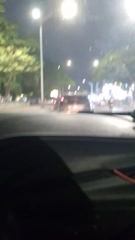 The video shows a car stopped at a traffic light at night. The view is from inside the car, looking through the windshield. Streetlights illuminate the road and surrounding trees. Several vehicles are visible in front, including a silver car with its brake lights on. A sign indicating "Zona Electronica Vigilada" can be seen. The interior of the car is partially visible on the right side of the frame, including the side window and the dashboard.
